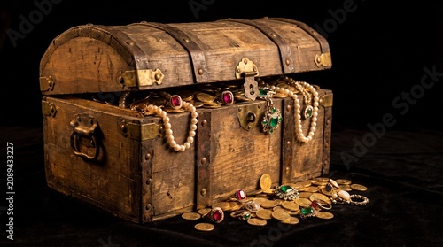 Antique wooden treasure chest filled with sparkling gold coins and precious gemstone jewelry overflowing on a dark black velvet surface