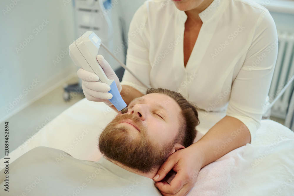 Fototapeta premium Close up of Caucasian young adult man receiving facial skin treatment from female clinician, targeting beard area for grooming and aesthetic care. Ideal for mens skincare marketing