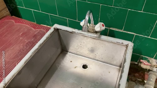 Rusty iron foot sink under faucet, set against green ceramic tiles, exposed pipes and valve, stained basin floor with clogged