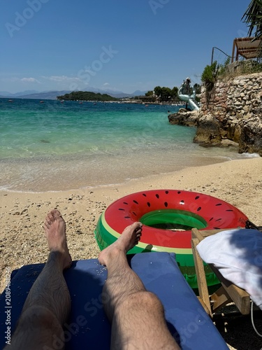 First-person view of a man relaxing on a sunny sandy beach sunbed near crystal-clear turquoise water on the Albanian coast, with a colorful watermelon-shaped inflatable ring in the foreground