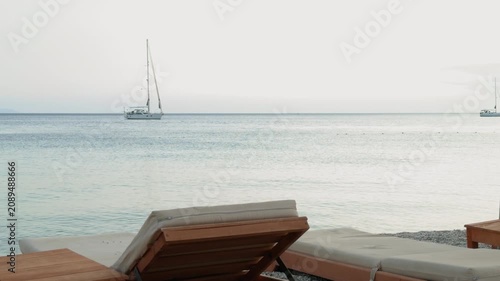 Comfortable wooden sun loungers overlooking a calm sea at sunset, with anchored sailboats on the horizon and soft pastel evening light  on the Albanian coast