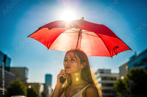 Exhausted young woman holding a red umbrella to protect herself from the intense glaring sun on a hot summer day in the city