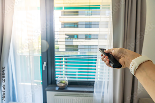 Woman controlling exterior window blinds with a remote control inside a modern apartment. Smart home automation and indoor climate control concept.