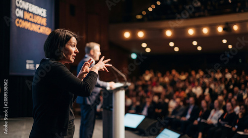 Sign language interpreter speaks beside conference presenter before audience for inclusive communication accessibility event professional education public lecture diversity inclusion