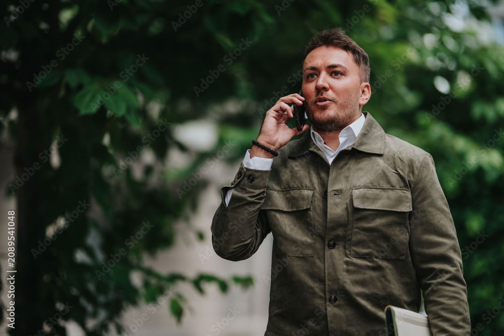 Fototapeta premium A businessman talks on his smartphone while holding a folder and walking outdoors. He pauses on an urban street, speaking on the phone while moving between destinations.