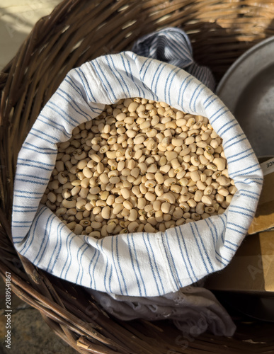 Rare Cretan white-eyed beans in a striped fabric sack inside a rustic wicker basket. Traditional Greek agricultural produce, vertical and high angle shot of raw organic local legumes.