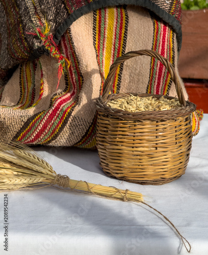 A small woven wicker basket filled with raw barley grains next to a tied wheat sheaf and a traditional striped Cretan textile sack on a rustic white table setting.