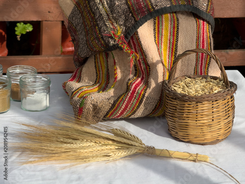 Traditional Greek harvest display with a wheat sheaf, a small wicker basket of barley grains, small spice jars, and a rustic woven textile sack on a white tablecloth.