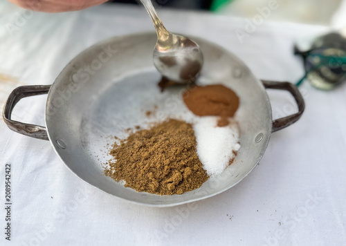 Measuring ground spices and white sugar with a spoon in a vintage metal pan. Preparation of traditional Cretan culinary recipes on a rustic white table setting.