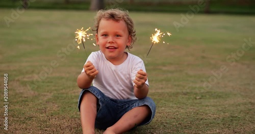 Young child smiles joyfully while sitting and holding glowing sparkler stick in each hand. Portrait shot captures his face glowing with delight and simple joy of childhood, as he celebrates something