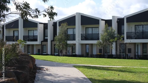 Modern two storey townhouses with dark roofline and balconies overlooking a footpath and lawn in an public green space in outer suburban Melbourne Australia. Contemporary medium density housing
