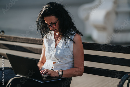 Obraz na plátně A woman in glasses types on a laptop while seated on a wooden bench