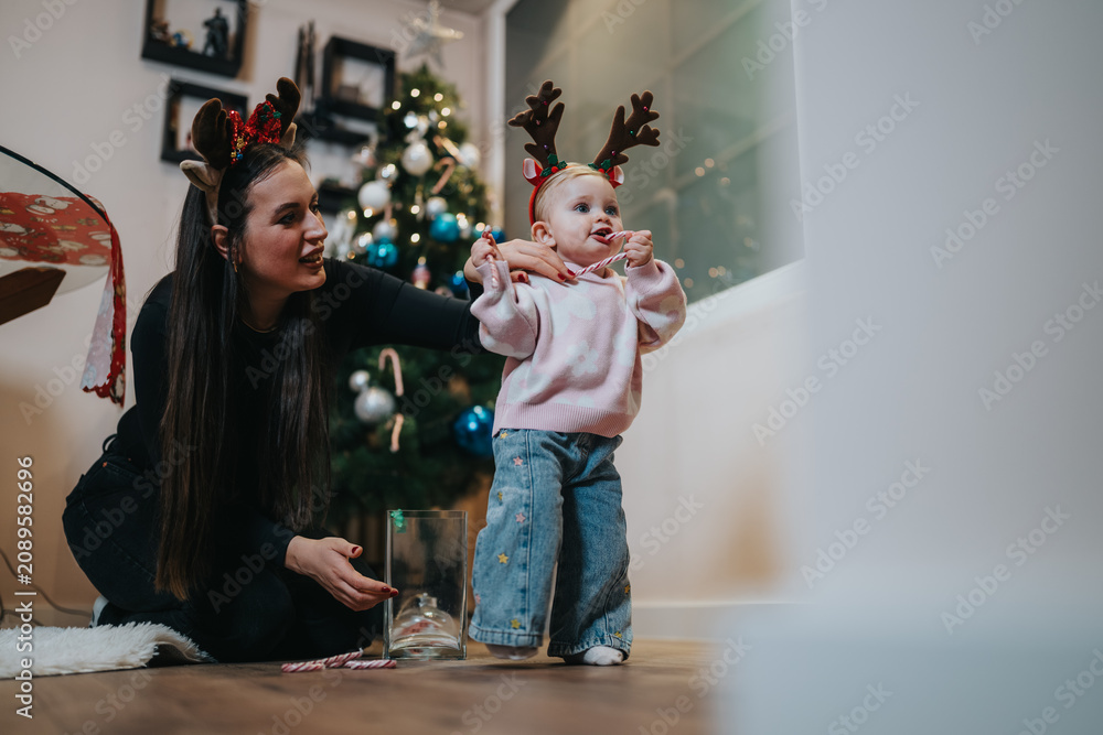 Fototapeta premium A mother helps her young child, both wearing holiday antler headbands, in a warm Christmas moment. They stand by a glowing tree inside a cozy home, creating a joyful family scene.
