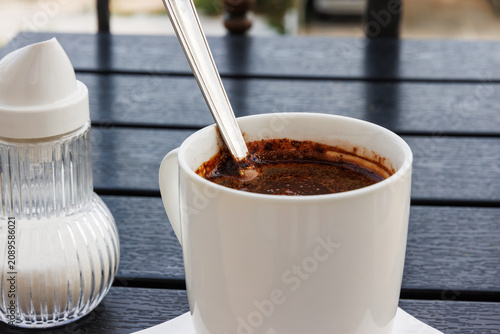 Closeup of black coffee in a white cup with spoon on a dark wooden table, with a sugar dispenser in the background.