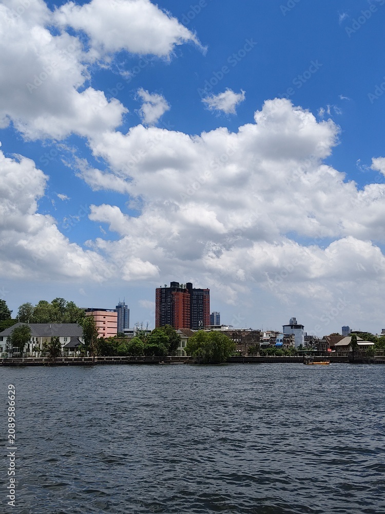 Fototapeta premium Scenic view of the Chao Phraya River with the Bangkok city skyline and a bright blue sky filled with white clouds.