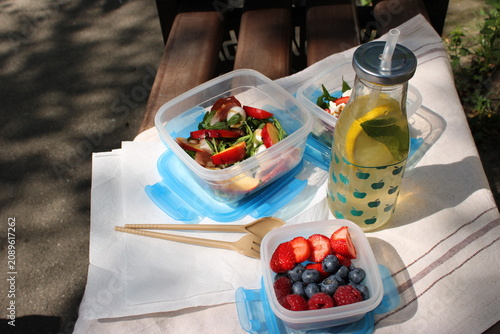 Salad and mixed berries in clear containers alongside a bottle of lemon water on a picnic blanket in a park, with sunlight filtering through trees