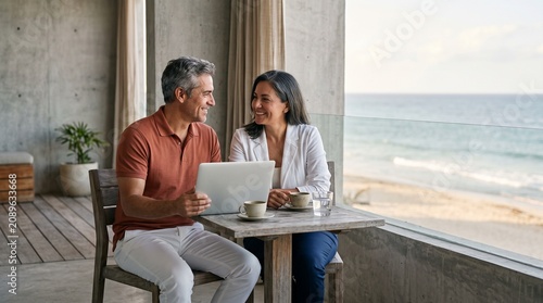 Senior couple smiling and collaborating with laptop on a sunny beach balcony