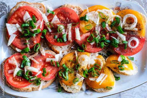 Close-up of fresh open-faced sandwiches topped with ripe sliced tomatoes, chopped white onion, green onions, black pepper, and a drizzle of oil on rustic bread. Traditional homemade summer snack 