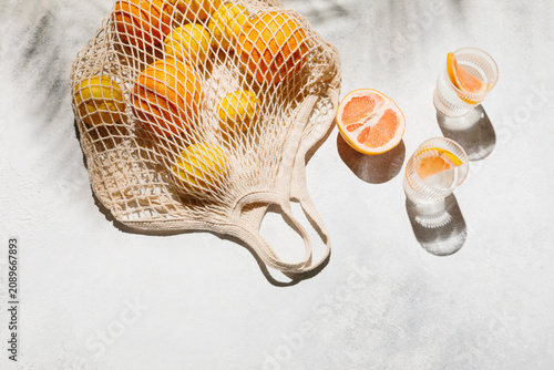 Fresh oranges and lemons in a mesh bag on a light surface with citrus drinks under bright sun. healthy summer lifestyle, zero waste shopping, tropical vacation spirit.