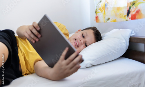Relax, smile and kid with tablet in bedroom for game, streaming show or learning online in home. Caucasian boy, bed or happy with digital technology for movie app, cartoon or reading ebook on weekend.