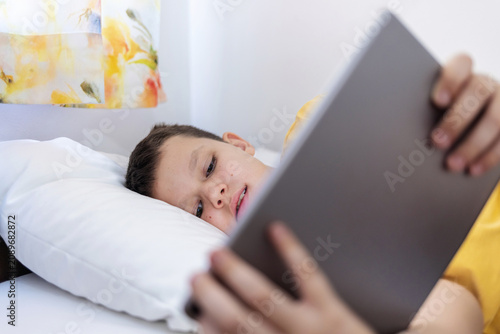 Boy lying in bed playing games on digital tablet at home. Happy little boy watching cartoons on digital tablet.