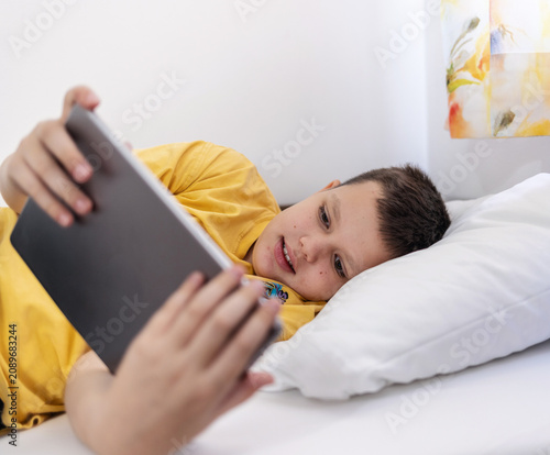 Boy lying in bed playing games on digital tablet at home. Happy little boy watching cartoons on digital tablet.