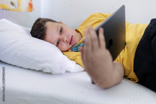 Young boy laying in bed and watching video clips on tablet. Boy lying on bed with digital tablet.