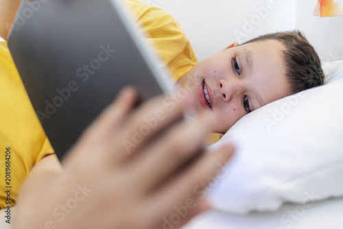 Young boy laying in bed and watching video clips on tablet. Boy lying on bed with digital tablet.