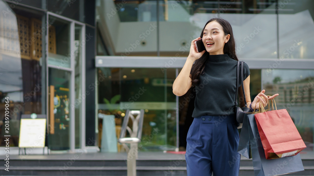 Fototapeta premium Asian woman talking on phone with shopping bags at modern mall, Black Friday, shopping