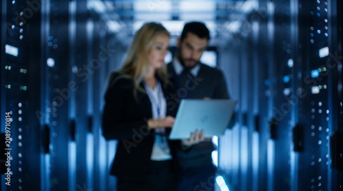 Professional data center technicians collaborating on server maintenance using laptop in modern high tech facility with blue ambient lighting and blurred server racks in background