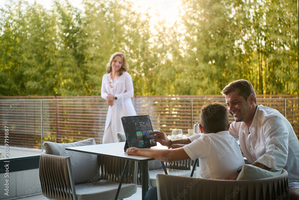 Fototapeta premium Happy Family on Modern Home Balcony Enjoying Sunset Together While Father Shows Son a Tablet