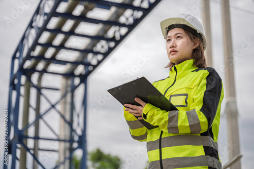 Professional female industrial engineer wearing safety gear uses a clipboard to inspect pipelines at a modern chemical plant facility to ensure reliable daily operations and compliance.