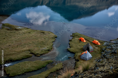 a small cluster of orange and grey expedition tents occupies a narrow tongue of vivid green moss beside a glacial stream where it spills from Carhuacocha lagoon into the valley below. 