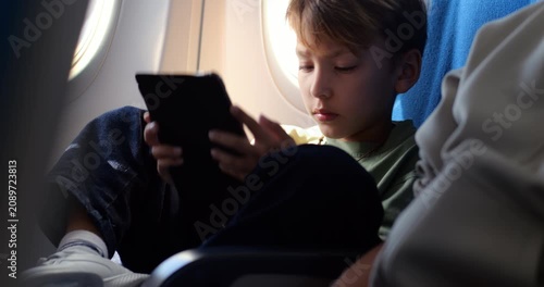 Young passenger reading digital book on his tablet while sitting near airplane window with legs crossed up on seat during flight. Boy enjoys quiet entertainment and peaceful journey