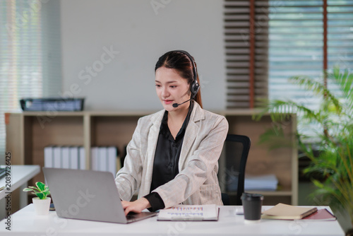 Young Asian woman wearing a headset and formal blazer typing on a laptop, providing professional customer support and telemarketing services from an office desk
