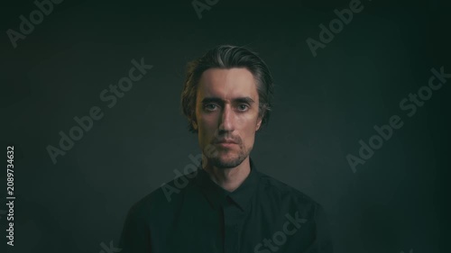 Young adult man seated against dark background in dark button-up shirt, low-key moody lighting highlighting facial features, introspective portrait, studio shot