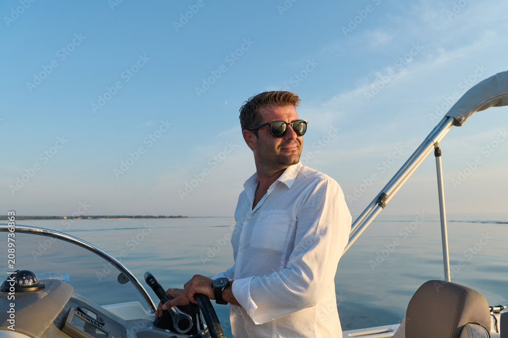 Fototapeta premium Man Steering Motorboat Near Umag At Sunset Wearing Sunglasses And White Shirt On Calm Sea