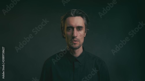 Moody studio portrait of adult man with medium-length dark hair and goatee in black collared shirt, looking at camera against dark gradient background