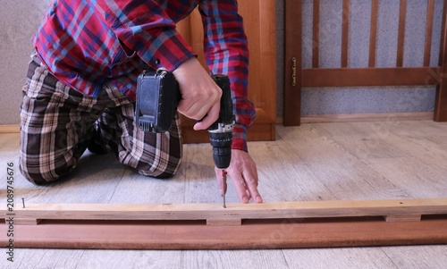 A man in a brightly colored plaid shirt kneels on the floor, screwing a screw into a wooden slat with a drill, assembling a bed. A room and brown wooden furniture are visible in the background.