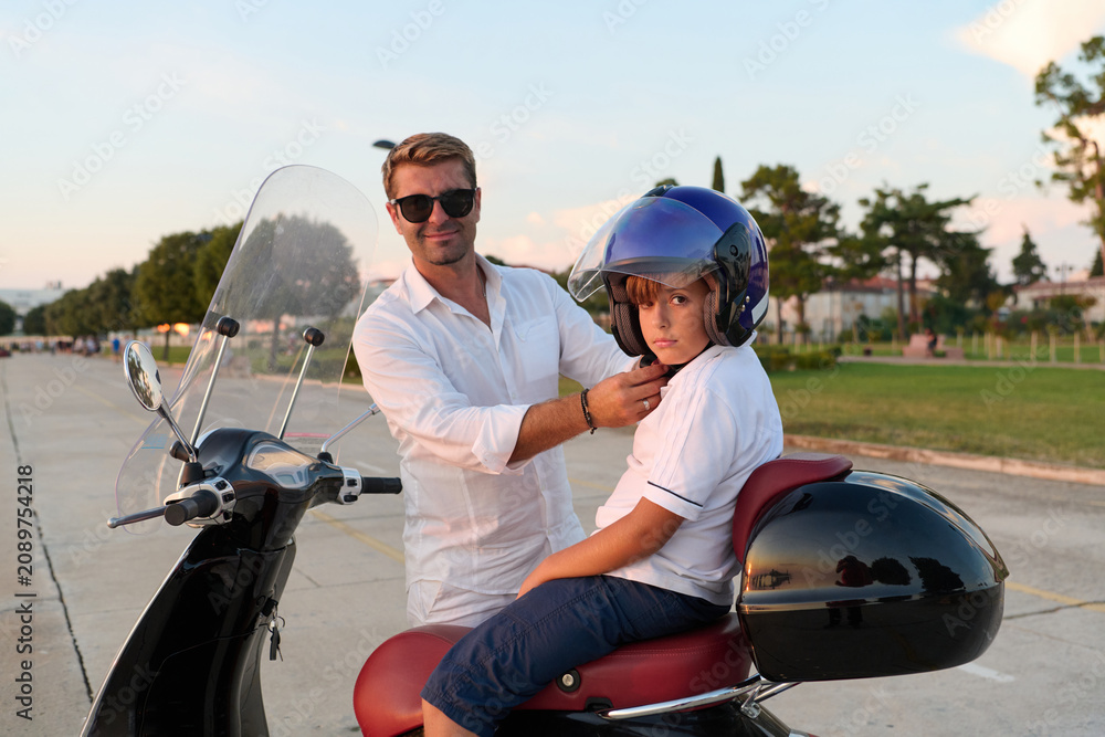 Naklejka premium Father Helping Son Fasten Helmet On Scooter During Summer Family Ride In Park