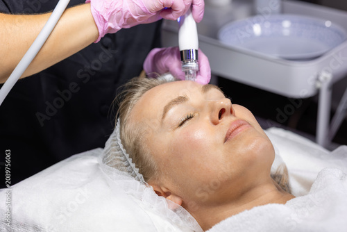 Cosmetologist performs hydradermabrasion on a woman's forehead using a hydro facial device to cleanse pores, exfoliate skin and improve hydration in a beauty clinic.