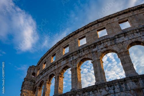 Canvastavla Pula, Croatia - August 18, 2025: Ancient Roman amphitheater captured from a low angle, its stone arches glowing in warm sunlight beneath a vivid blue sky