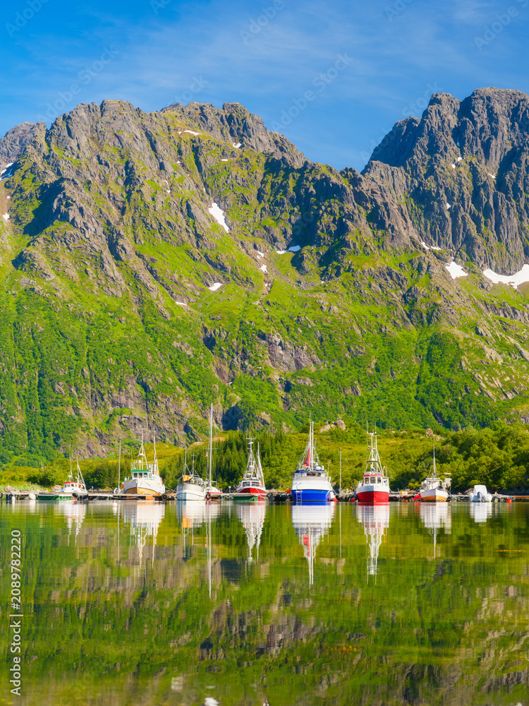 Fototapeta premium Fishing boats and ships anchored in a sea bay at Lofoten islands, Norway. Vessels reflecting on the calm water surface during summer.