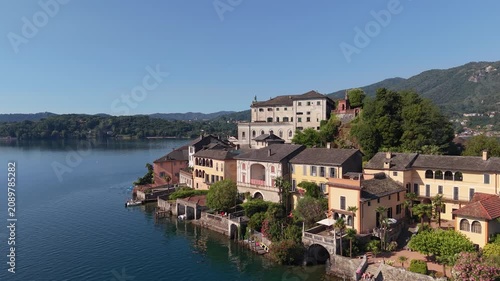 Aerial drone orbit view of San Giulio Island on Lake Orta, showing the historic lakeside village, ancient stone buildings, basilica tower, monastery complex, clear blue water, and surrounding hills. S