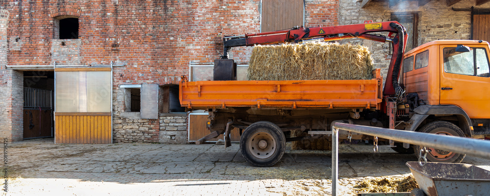 Fototapeta premium Orange farm truck with crane carries hay bales beside brick stable yard, showing rural work and equestrian life. Sunlight, straw, stone walls and barn doors create authentic countryside atmosphere