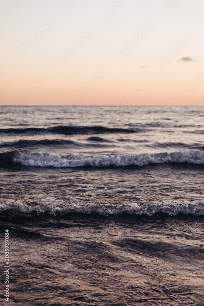 Fototapeta premium Close-up of dark, moody ocean waves rolling and breaking near the shore. Textured water surface and sea foam reflect soft, warm light of twilight, creating a dramatic and abstract marine background.