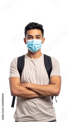 A young man with dark hair wearing a blue medical face mask and a tan t-shirt stands with his arms crossed
