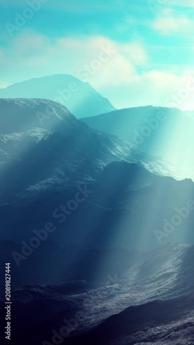 Light beams shine through a mountain range during early morning. The scene shows layers of mountains with a hazy atmosphere and soft light illuminating the landscape.