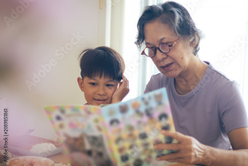 Canvastavla An Asian grandmother wearing eyeglasses reading an illustrated book with her cute young grandson at the table, sharing an educational and heartwarming family bonding moment inside a bright room