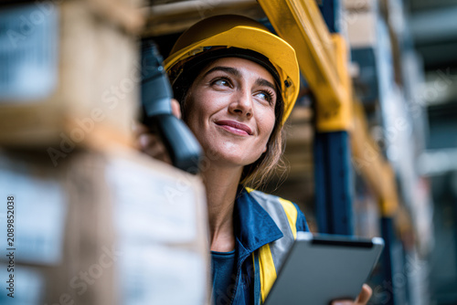 A female worker wearing a yellow hard hat and safety vest holds a digital tablet while inspecting inventory in a warehouse filled with stacked boxes.